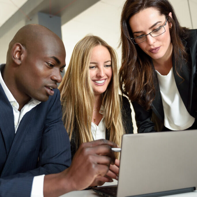 Multi-ethnic group of three businesspeople meeting in a modern office. Two caucasian women and a black man wearing suit looking at a laptop computer.
